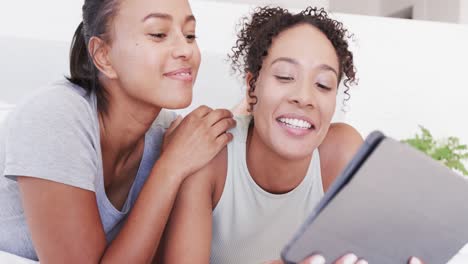 Happy-biracial-lesbian-couple-lying-on-bed-using-tablet,-in-slow-motion