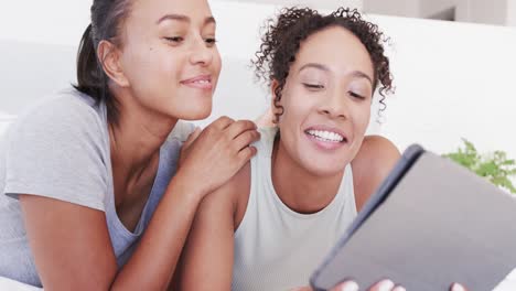 una feliz pareja lesbiana biracial acostada en la cama usando una tableta, en cámara lenta