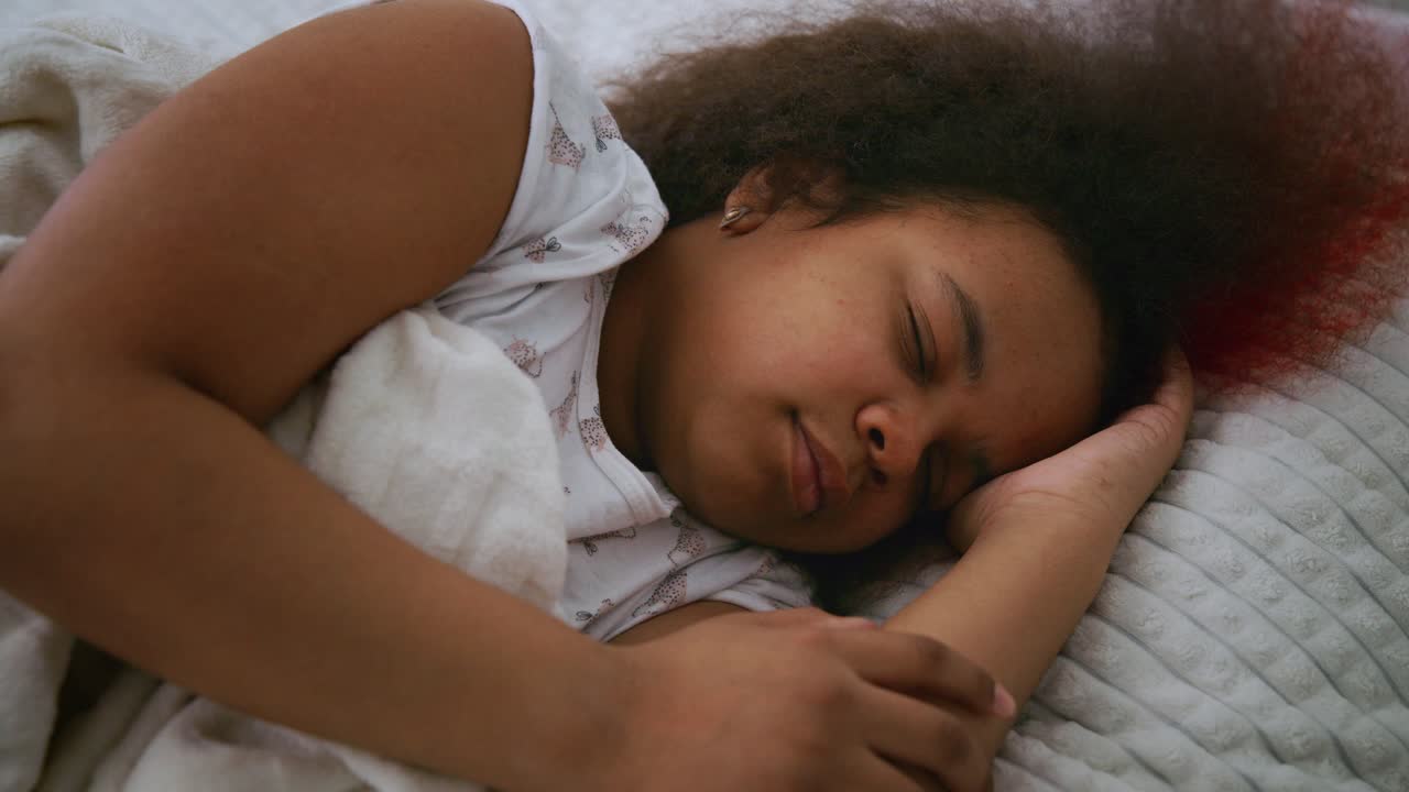 Young african american girl with partially red curly hair drifting peacefully in deep sleep, gently changing positions while nestled comfortably under soft blankets in quiet bedroom