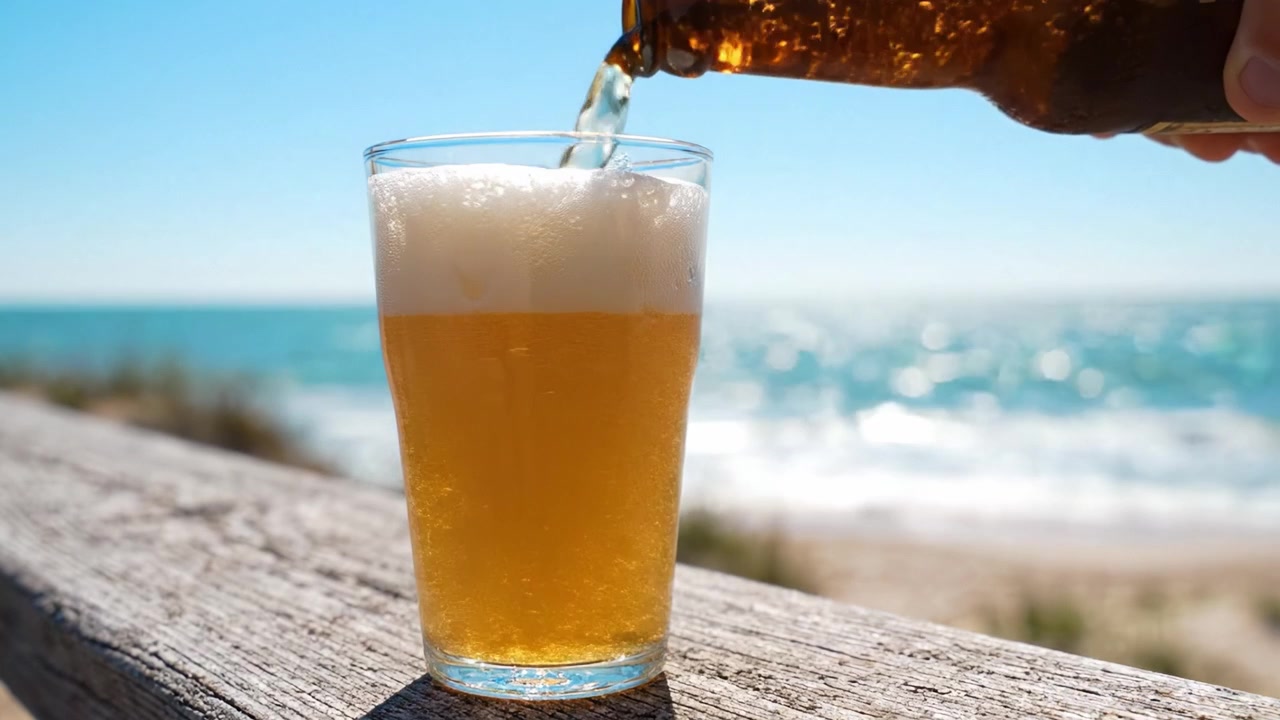 Momento rinfrescante in spiaggia: versare birra ghiacciata in un bicchiere contro uno splendido sfondo oceanico con onde scintillanti sotto il cielo azzurro e limpido.