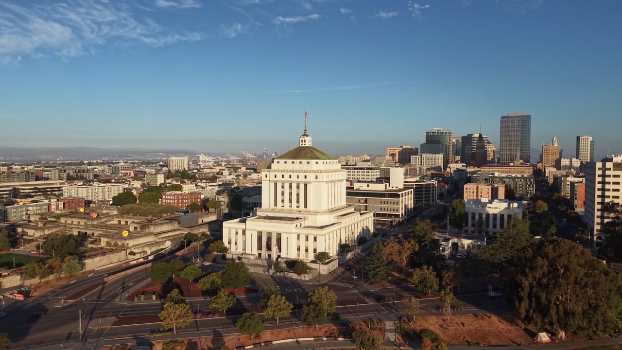 Luce solare mattutina che illumina il tribunale superiore della contea di Alameda con lo skyline del centro di Oakland, California, e il porto di Oakland visibili sullo sfondo sotto un cielo azzurro e limpido