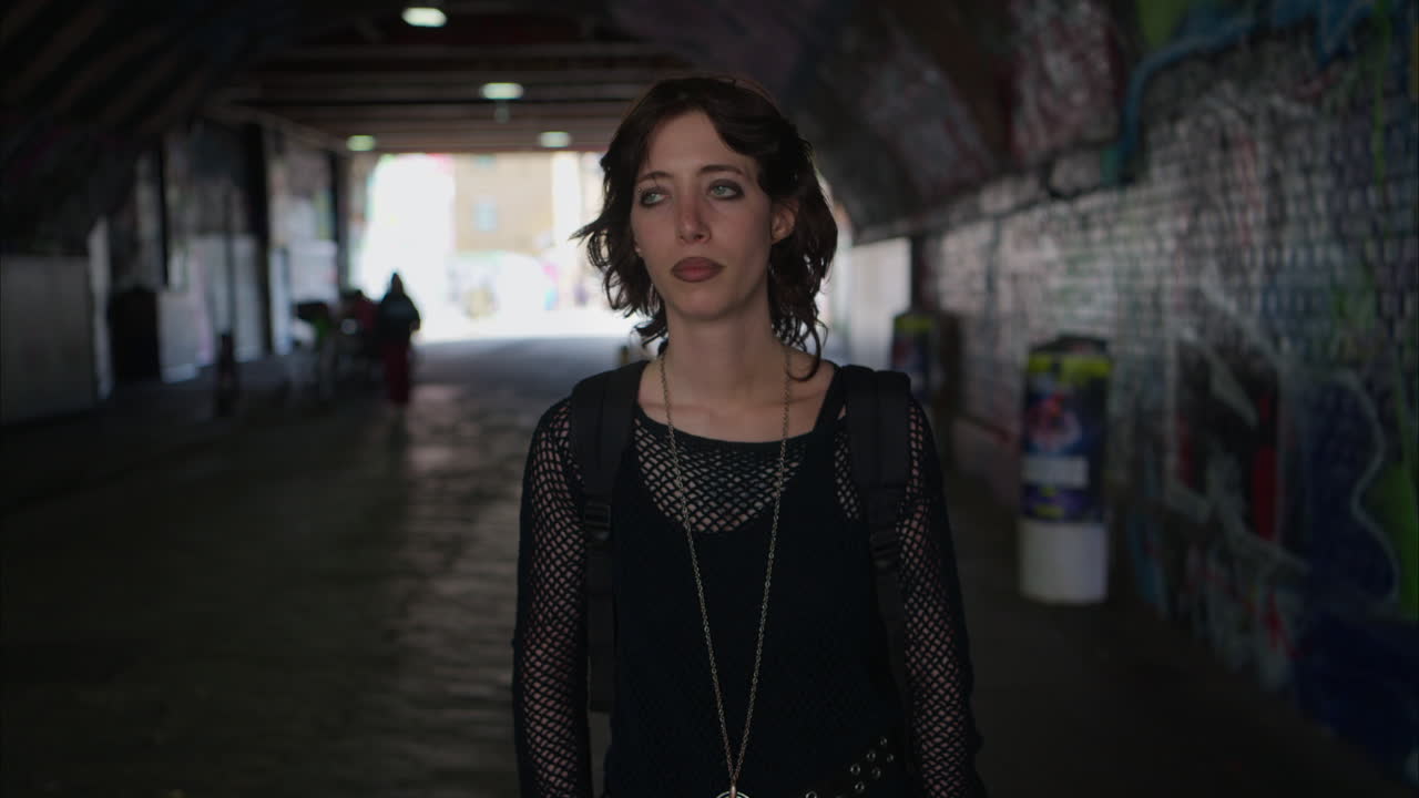 retrato de moda de una mujer joven de estilo alternativo caminando a lo largo de un paso subterráneo oscuro con paredes cubiertas de graffiti en la calle de la ciudad de londres, reino unido, en tiempo real.