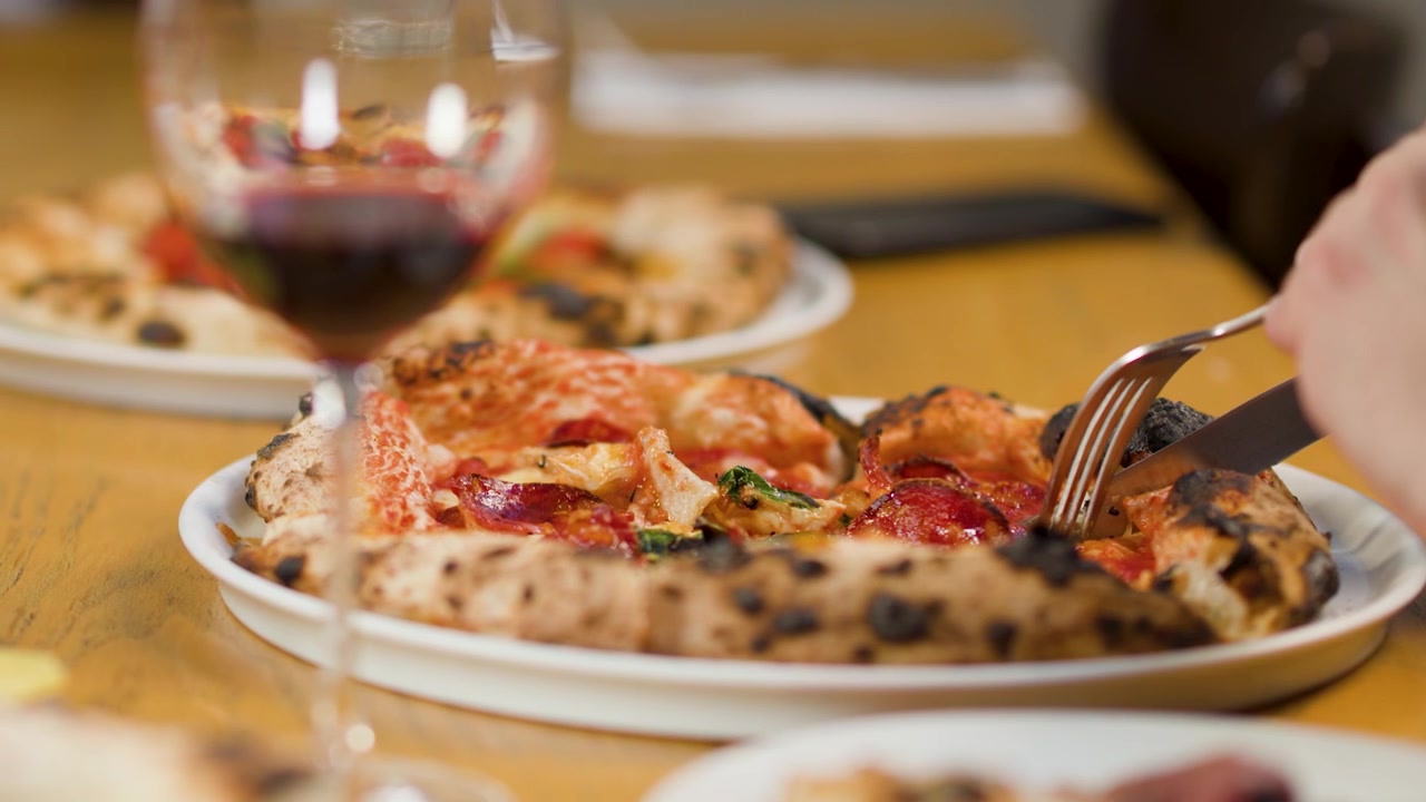 Mani che usano posate d'argento per tagliare una pizza fresca cotta nel forno a legna accanto a un bicchiere di vino rosso
