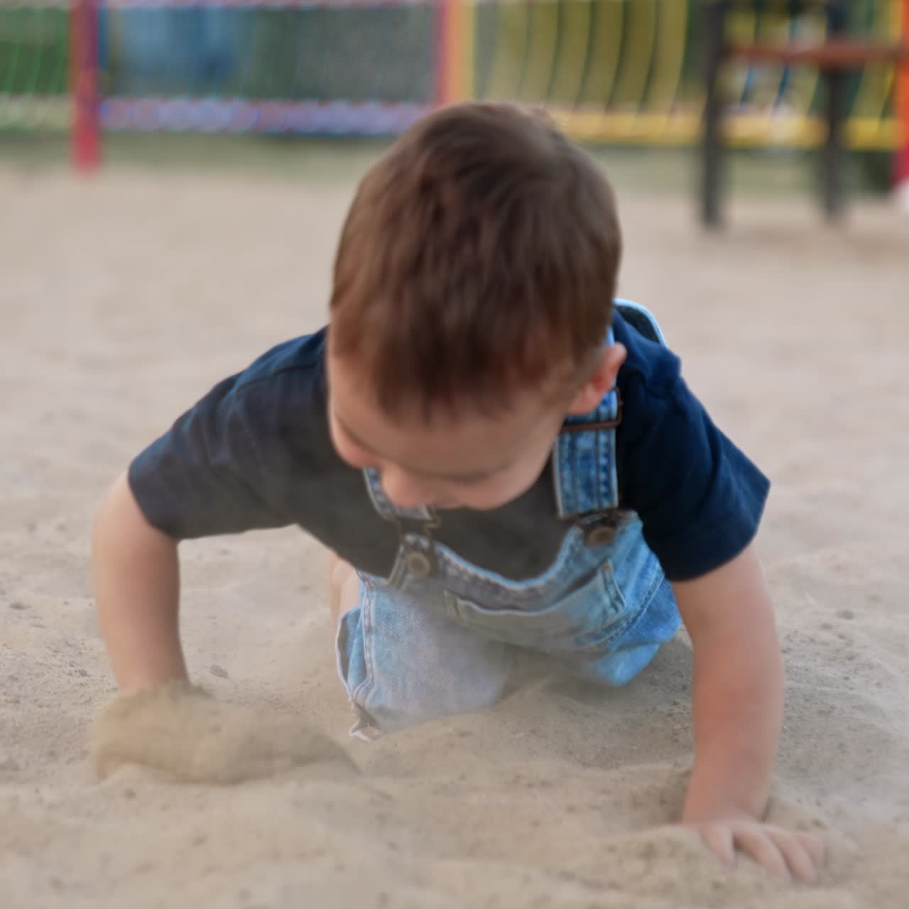 Active Caucasian toddler in jeans romper playing outdoors. Barefoot kid walks by the sand, then stands on his knees crawling by the sand