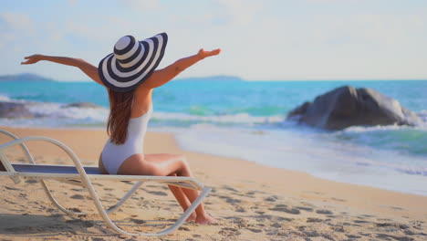 chica en forma irreconocible en traje de baño y sombrero a rayas sentada en una tumbona cerca del mar cuando el agua rueda sobre la playa de arena durante el día