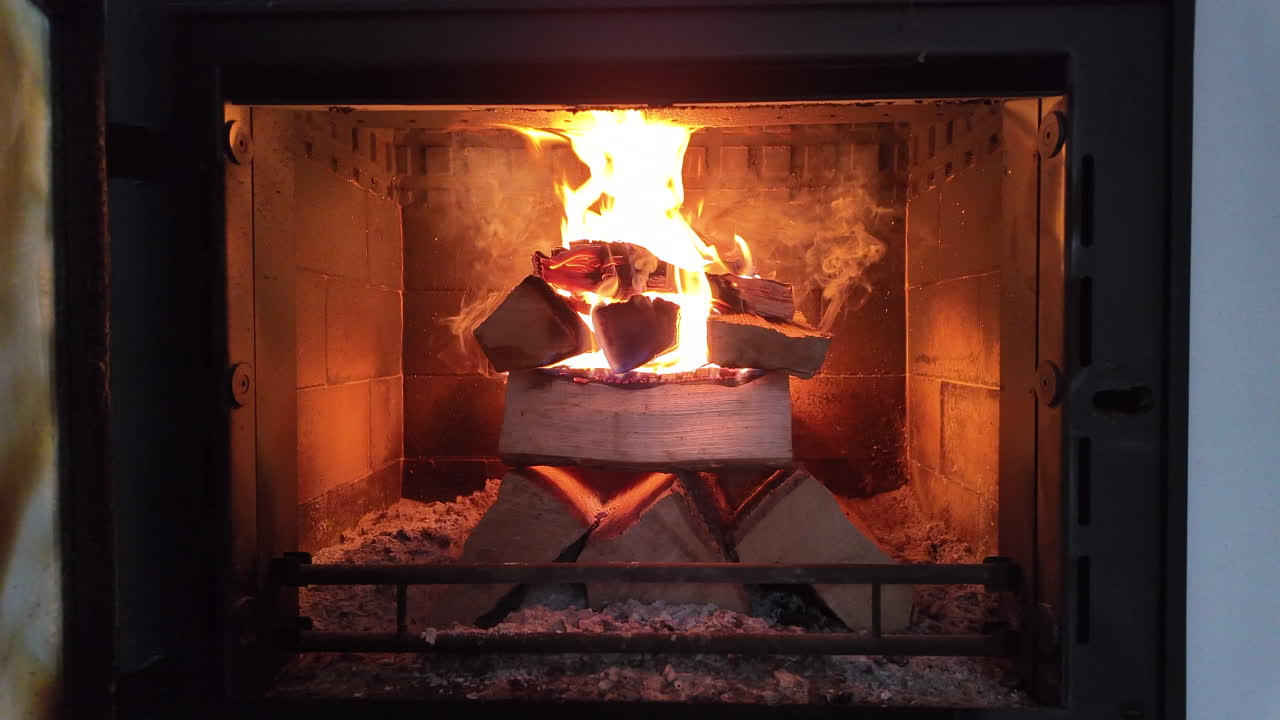 A bright, steady fire burns inside a modern enclosed fireplace, with stacked firewood glowing as tall flames rise in the center. The warm orange light reflects off the fireproof interior,