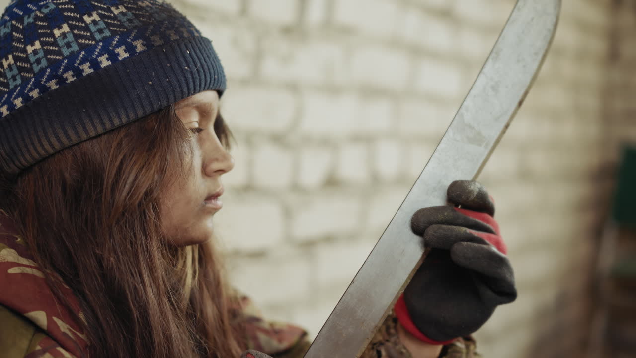 Dirty kid in wool cap and gloves holding cutlass thoughtfully near brick wall, face smeared with dust, gazing at blade with serious expression in worn jacket showing hardship and resilience