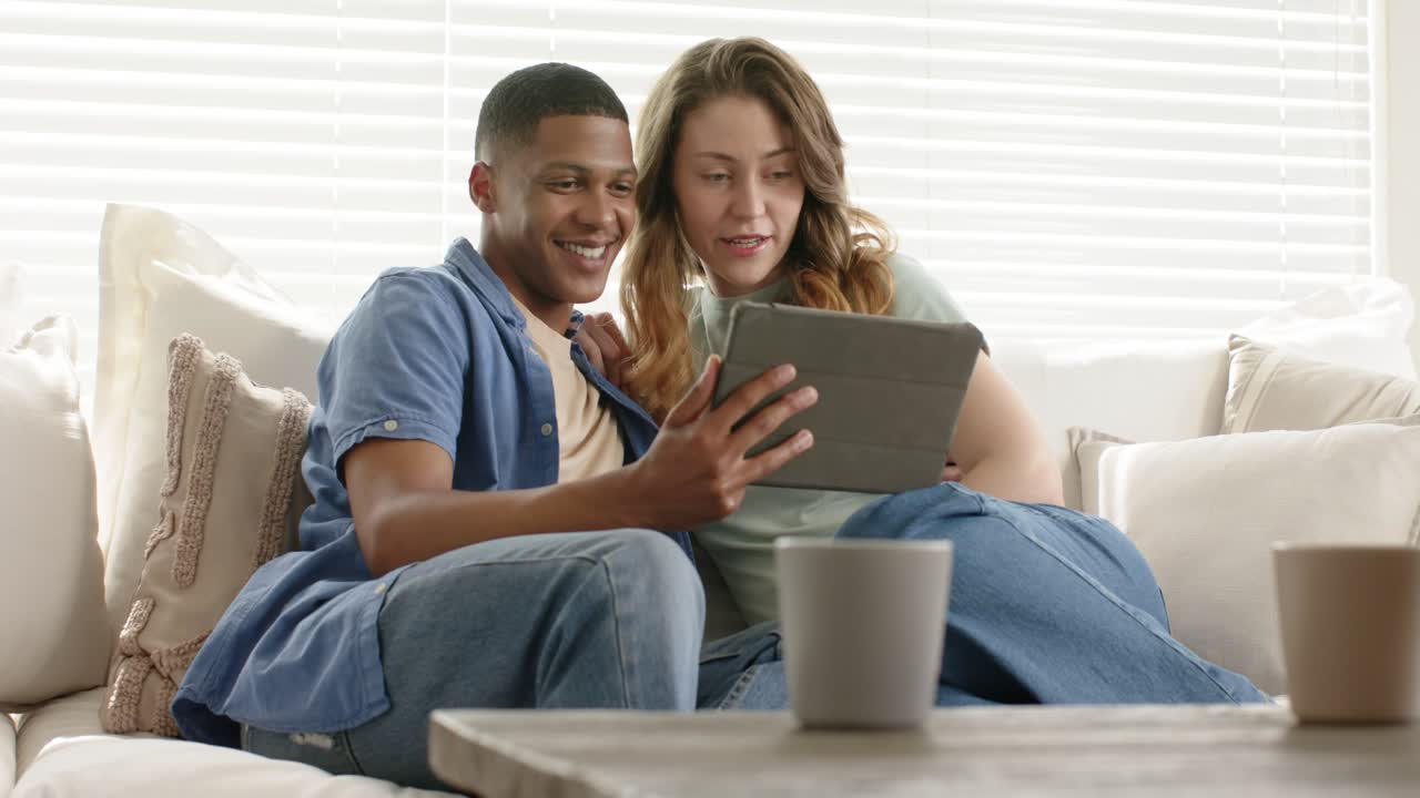 Viewing tablet screen, diverse couple tapping swiping sharing moment on sofa with mugs smiling