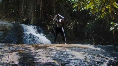 Hombre-Africano-Atlético-Sin-Camisa-Parado-En-Una-Roca-Junto-A-La-Cascada-En-El-Bosque-Tropical-Mirando-Hacia-Arriba---Alejar,-ángulo-Bajo