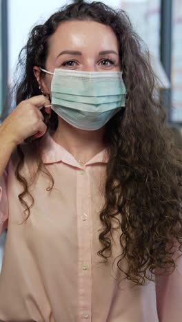 dark-haired woman wearing mask in office. girl showing her fingers at the mask, takes it off and smiles cheerfully. happy lady holding a mask in her hand by the face. vertical video