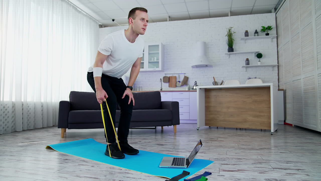 Concentrated man exercising with a rubber band at home. Young man doing working out while standing on a mat in the kitchen. Sportive lifestyle during quarantine.