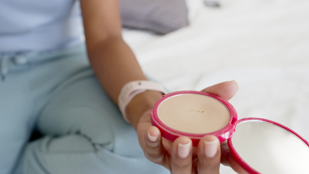 Brush is entering frame, tapping and sweeping pink compact to load bristles for makeup on bed