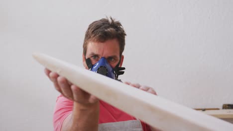 caucasian male surfboard maker wearing a breathing face mask and checking a wooden surfboard