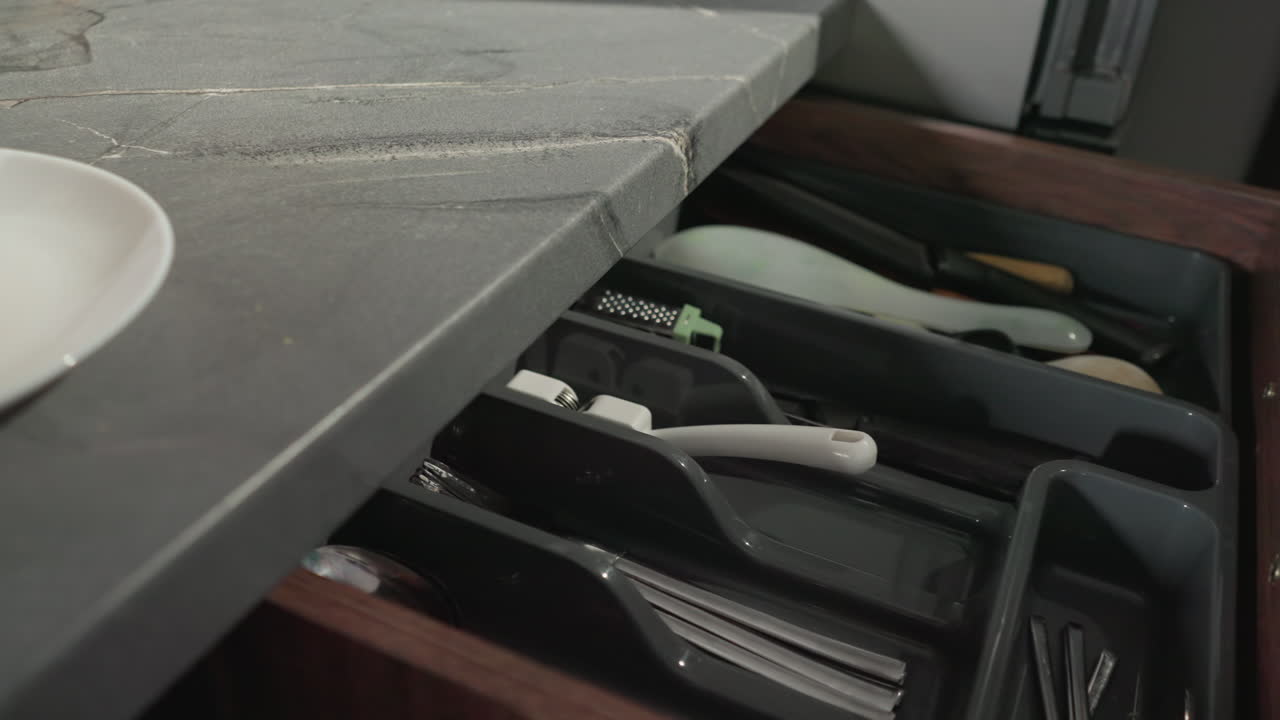 Older woman opening kitchen drawer to retrieve something in modern kitchen, with kitchen countertop, appliances, and utensils visible, focused on home cooking process in comfortable setting