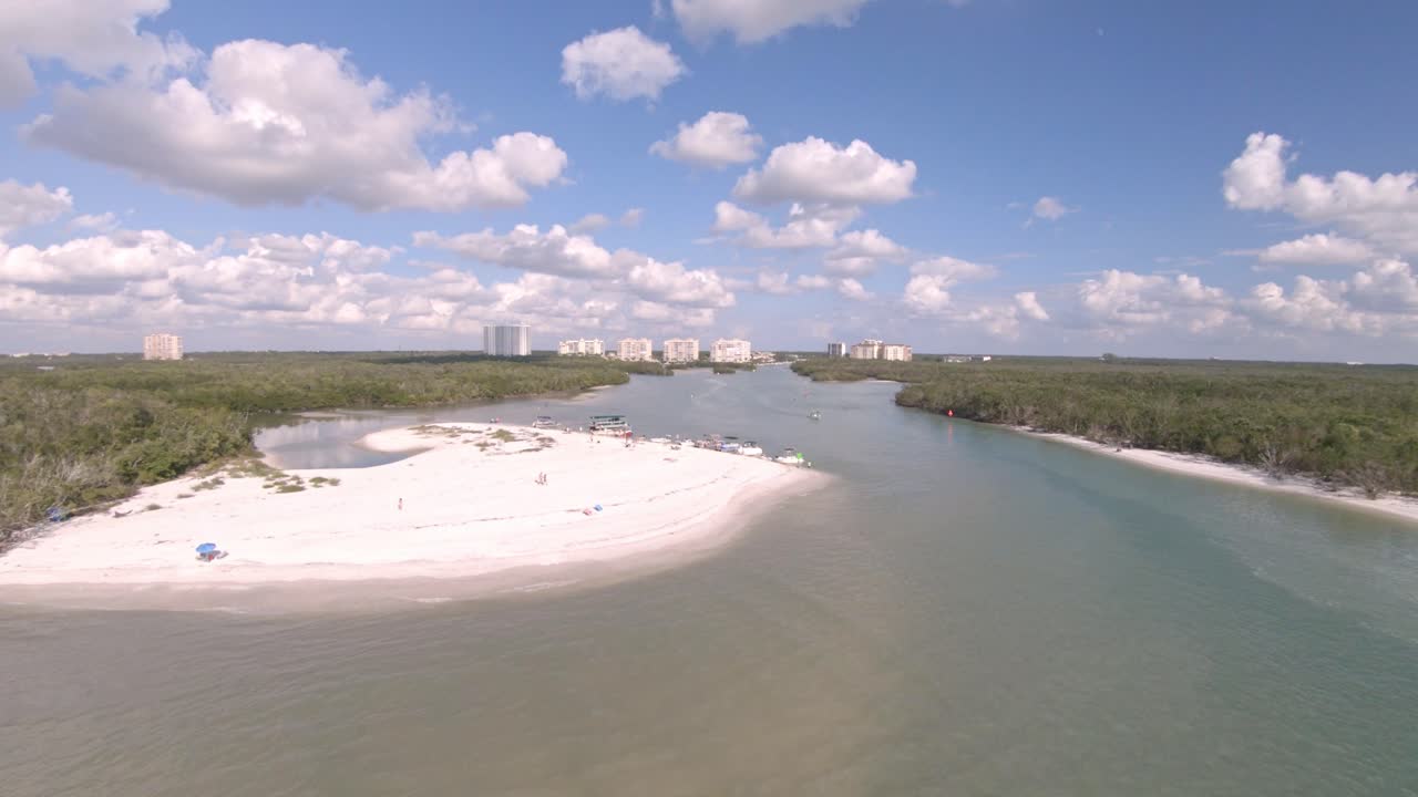 Drone video moves backwards over Wiggins Pass a beach river opening with boats moored on the beach. Trees surround and a residential community can be seen inland. Sunny, bright day with clouds above.