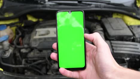 joven sostiene un teléfono inteligente móvil en la mano y da vueltas desliza a la derecha su dedo en la pantalla en el compartimento del motor de fondo del coche, coche con un capó abierto