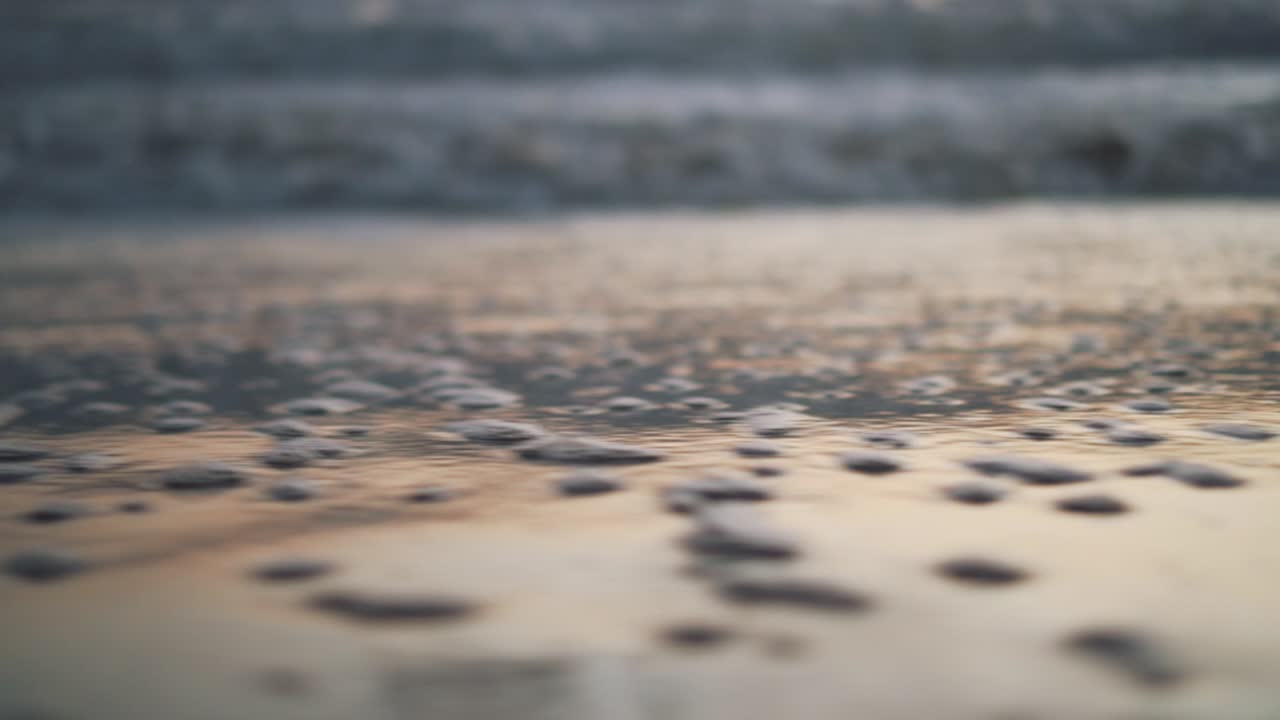 Close-up of the shoreline with water bubbles reflecting the colours of the sunset during golden hour - Trucking movement from left to right with blurry background