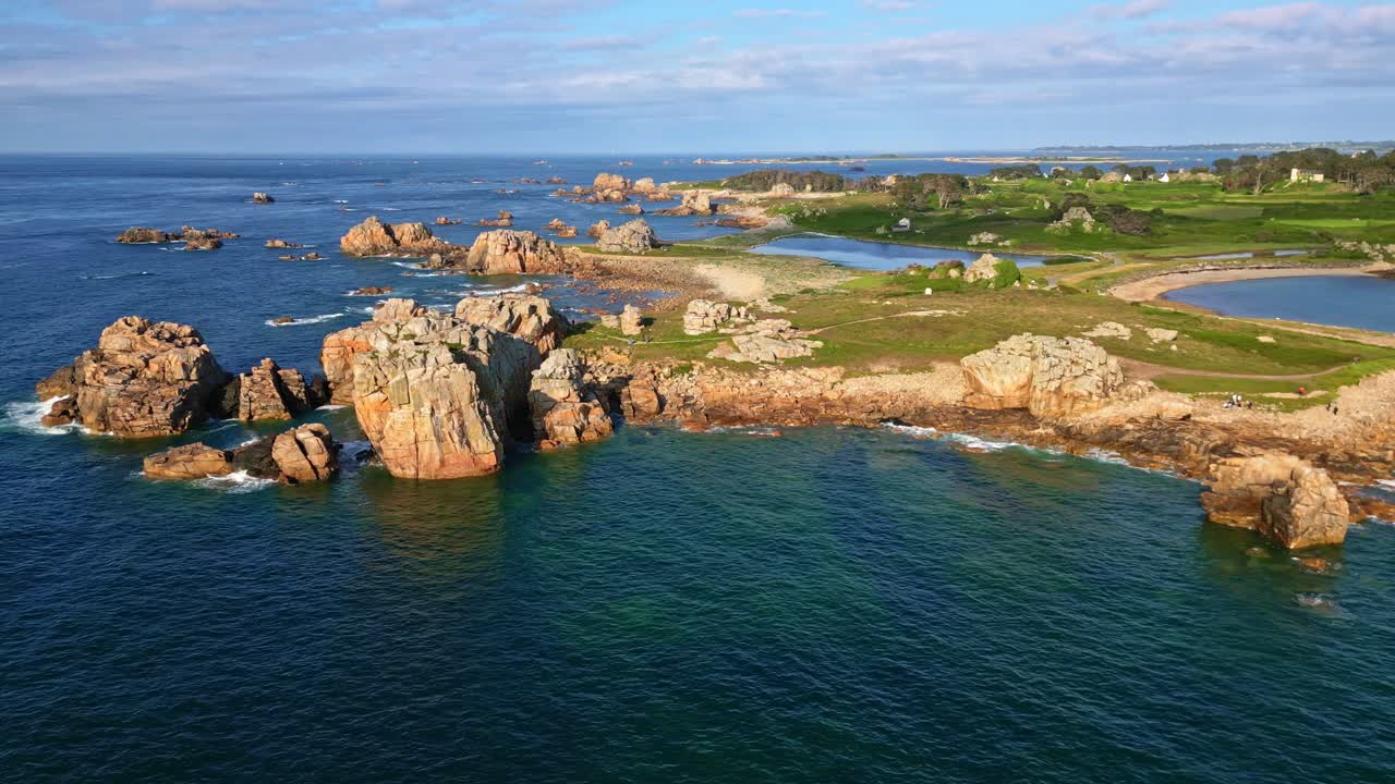 Drone shot over blue sea showing rocky coastline, hiking path, green vegetation, and distant shore at Gouffre de Plougrescant - Brittany France
