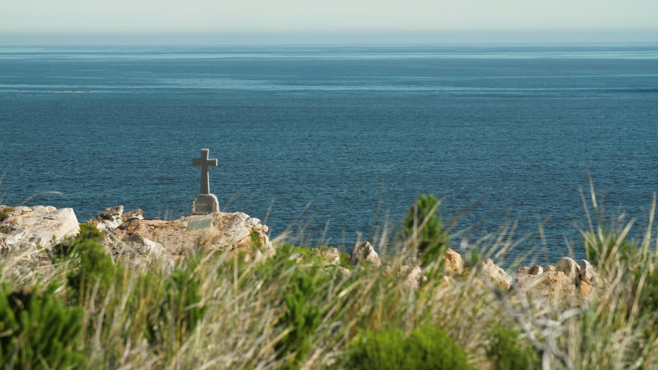 Coastal view of gravestone overlooking blue ocean with green grass - shrubs in foreground and heatwaves visible