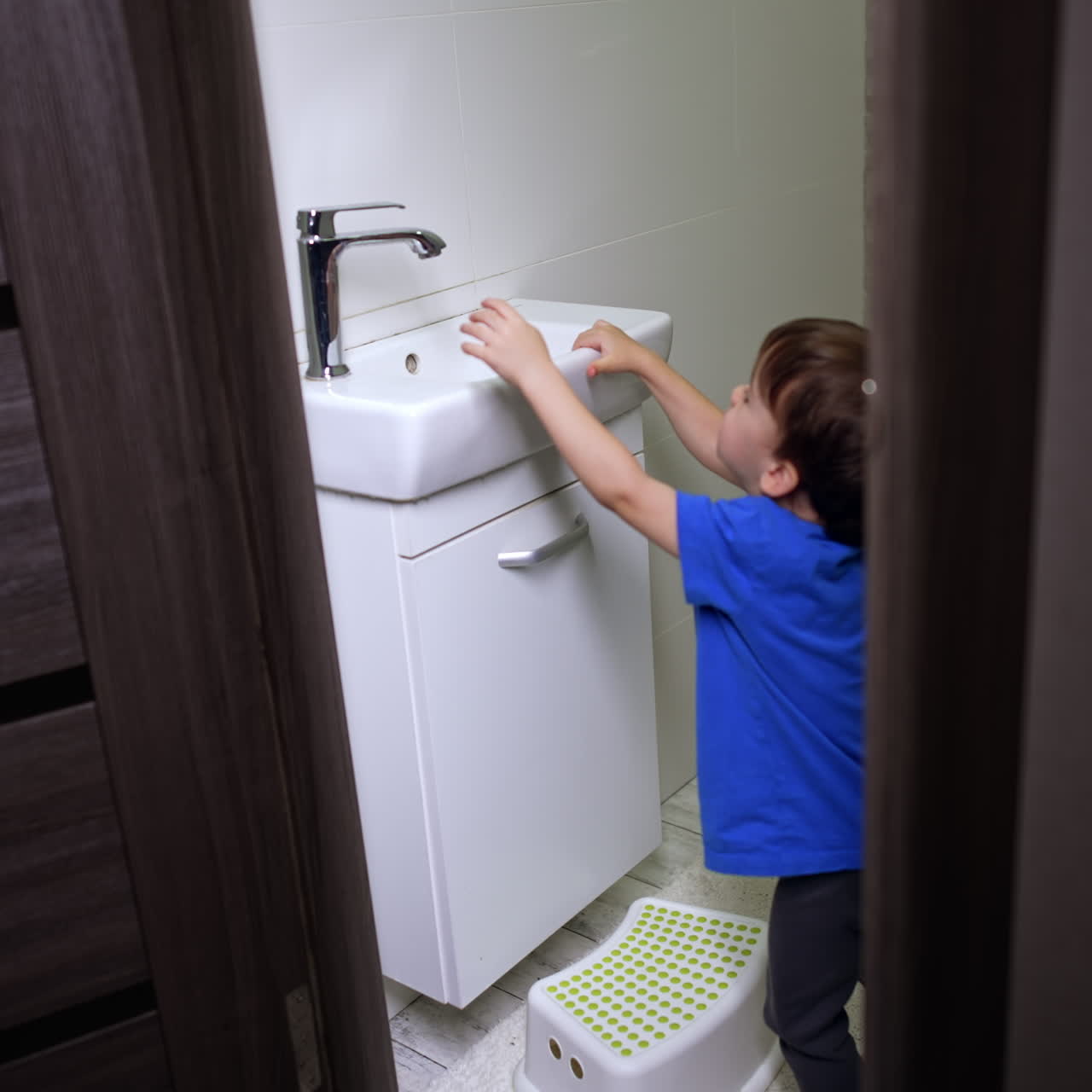 Peeping into the bathroom where little kid stands near the sink. Toddler opens and closes the tap, watching the water flow