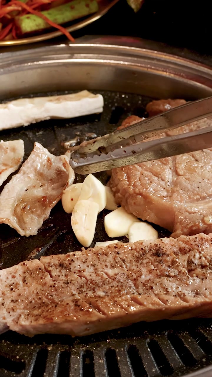Tongs move garlic cloves on sizzling pork belly slices over a tabletop Korean barbecue grill