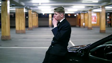 young man in a black suit with a bow-tie smoking a cigarette sitting at the bonnet of the black car in the parking