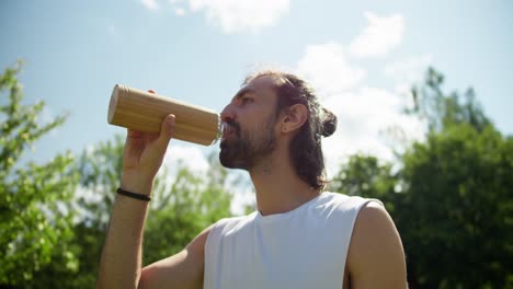 un tipo moreno con una camiseta blanca bebe agua de una botella amarilla especial contra el fondo de árboles verdes y un cielo azul