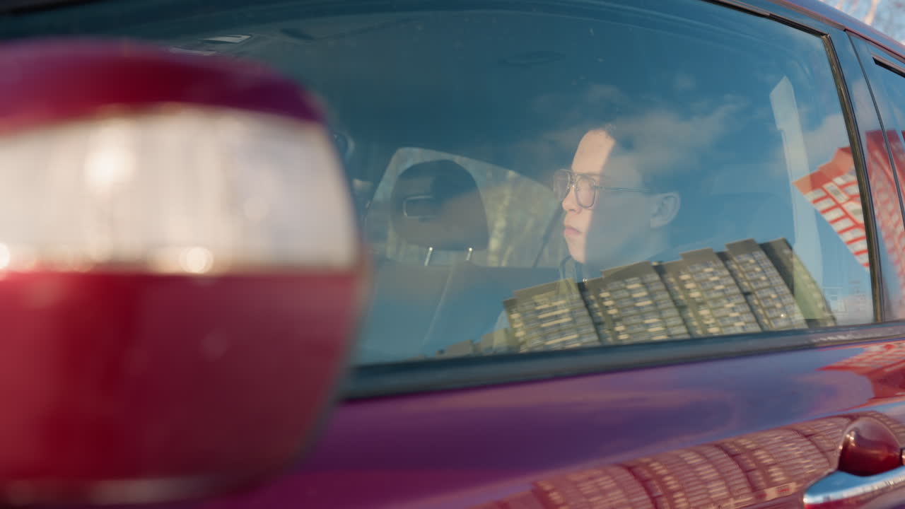 close up of red car window reflecting office building and student eating chocolate and sipping juice inside parked car with blurred winter urban backdrop conveying candid snack moment