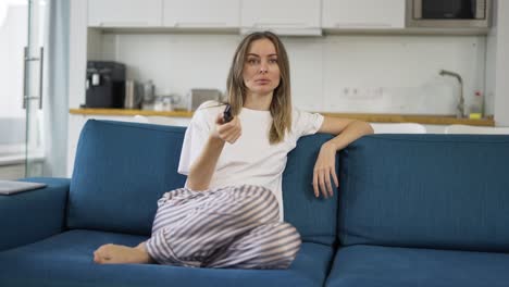 una mujer joven sentada en un sofá con un control remoto de tv y cambiando de canal, vista frontal