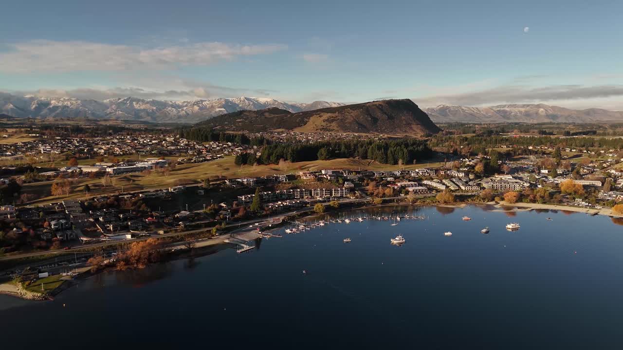 Aerial over Wanaka town harbor with boats on calm lake at sunset, with snow-capped Southern Alps in background, New Zealand