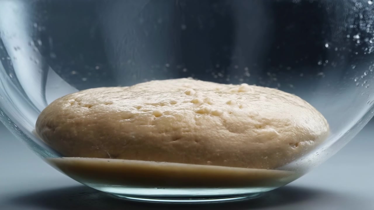 Low-angle close-up of proofing bread dough resting in clear glass mixing bowl, with flattened dome shape, moist pale beige surface, tiny pores, dimples, smooth elasticity, and soft studio reflections