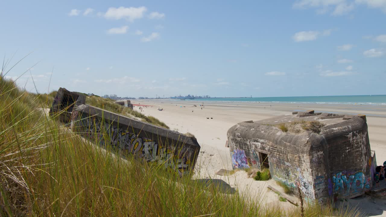 Static wide shot of graffiti-covered bunkers on sandy beach, bright daylight, calm seaside atmosphere