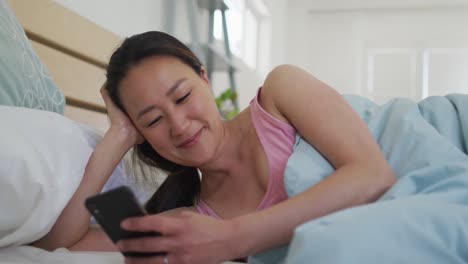 아침에 스마트폰을 사용하여 침대에 누워있는 행복한 아시아 여성