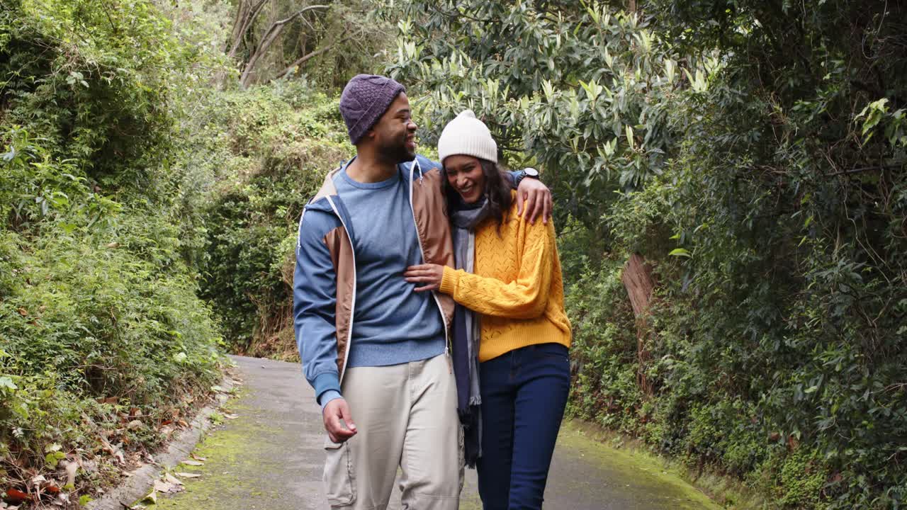 Diverse couple linking arms on paved path in park, man glancing causing woman smiling with scarf