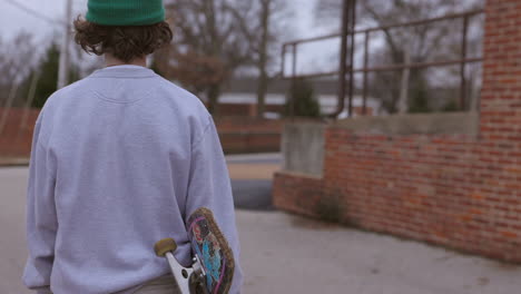 teen boy with a skateboard walks away and towards a back alley parking lot