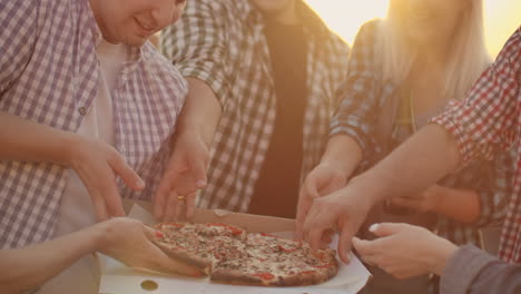 a company of six young people parses pieces of hot pizza and eats together. this is a rooftop party with a beer.