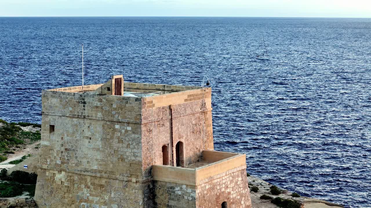 Et majestetisk vidvinkelbilde av det historiske Dwejra-tårnet som vokter over det dypblå Middelhavet på en lys sommerdag på Gozo, Malta, med varm, gyllen kalkstein mot en klar himmel