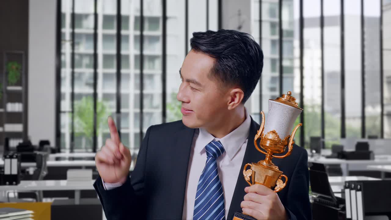 Close Up Of Asian Business Man In A Suit And Tie With A Gold Medal Holding A Gold Trophy In His Hands, Smiling, And Pointing To Side In Office