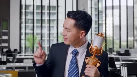 close up of asian business man in a suit and tie with a gold medal holding a gold trophy in his hands, smiling, and pointing to side in office