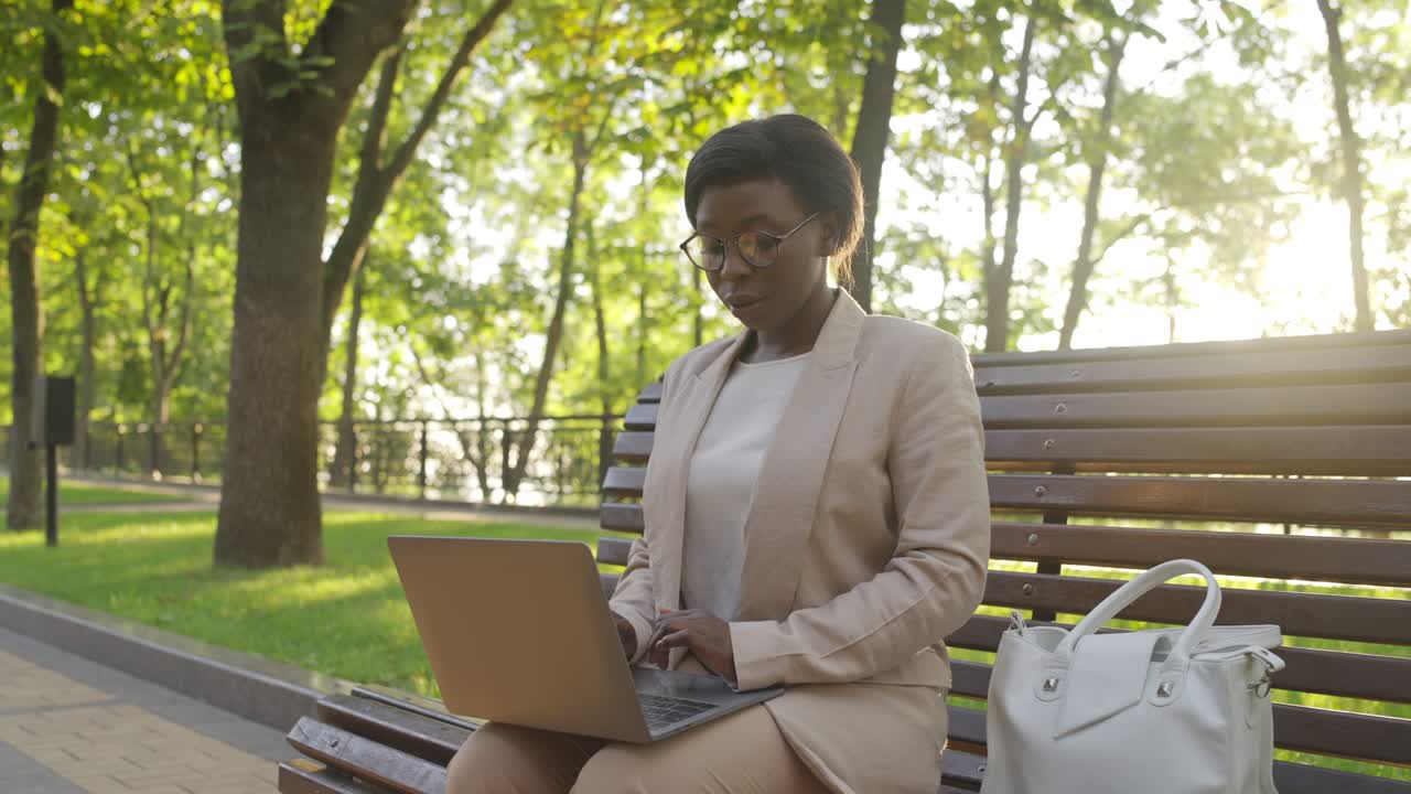 mujer afroamericana trabajando en una computadora portátil, sentada en un banco en un parque de verano