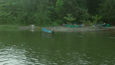 Blaues-Boot-Am-Flussufer-Mit-Grünen-Bäumen-Im-Hintergrund-Und-Einem-Vorbeigehenden-Hund