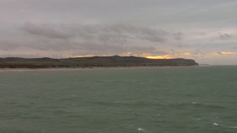 Vista-Costera-De-La-Costa-Francesa-Desde-El-Canal-De-La-Mancha-Al-Atardecer,-Con-Suaves-Olas-Y-Cielos-Nublados