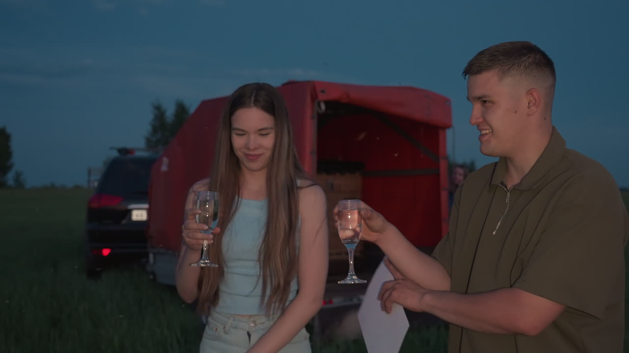 couple raising champagne glasses while holding certificate on grassy field at sunset reacts to buzzing insects flying around them by red balloon trailer and wicker basket