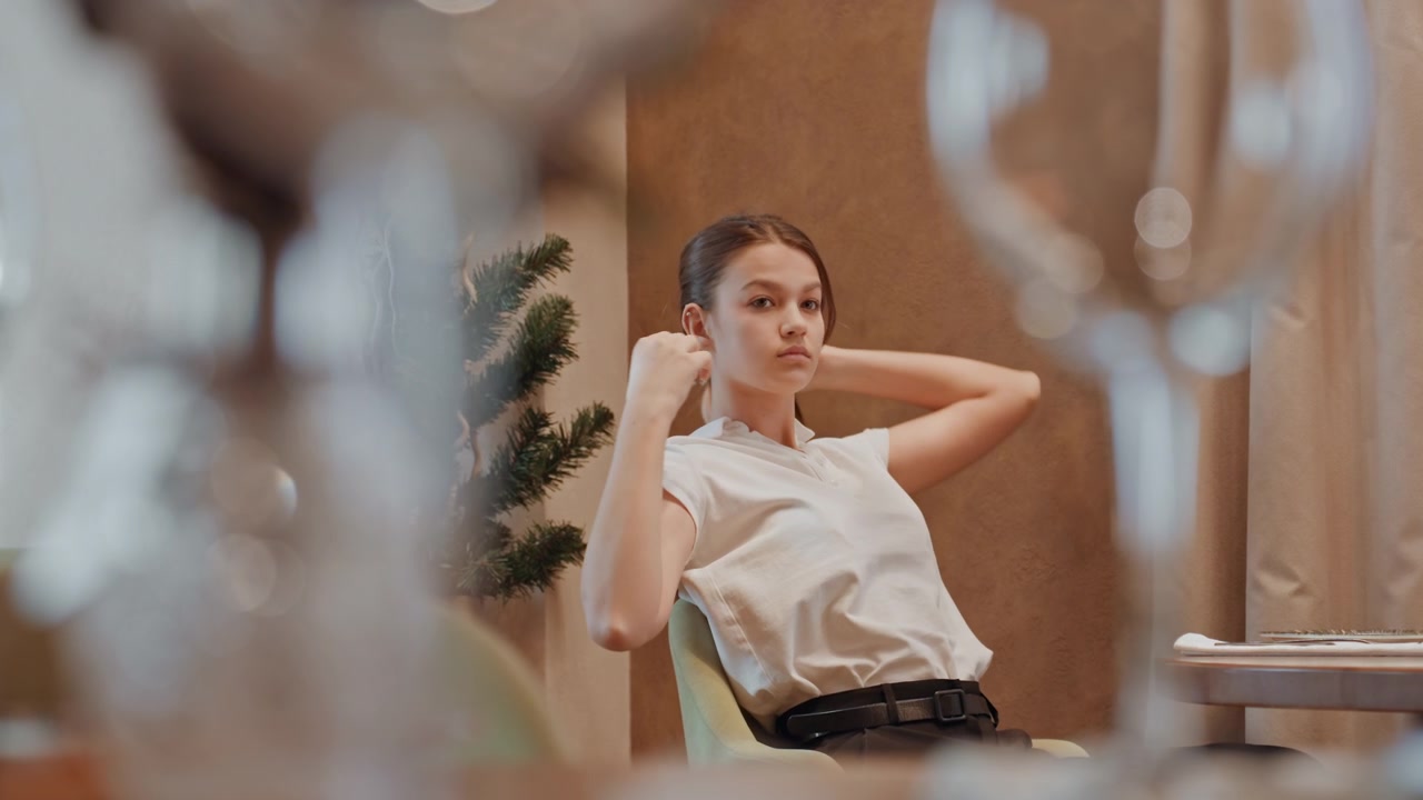 Young white woman in restaurant chair, styling hair through reflection, preparing for evening engagement, blurred glassware foreground, candid motion, tailored blouse, confident expression, editorial