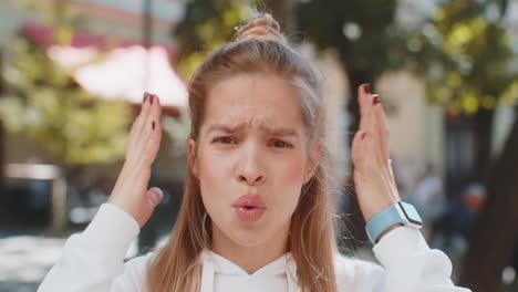 closeup portrait of shocked young woman teenager touching face and looking at camera on city street
