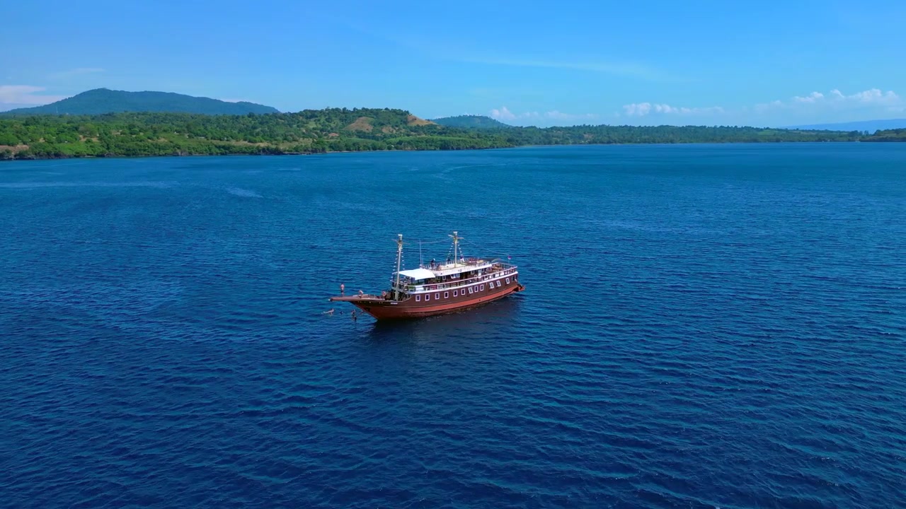 Perahu pinisi tradisional di perairan terbuka dengan seseorang melompat dari haluan ke laut. Pemandangan udara menakjubkan dengan panorama orbit lebar dari drone.