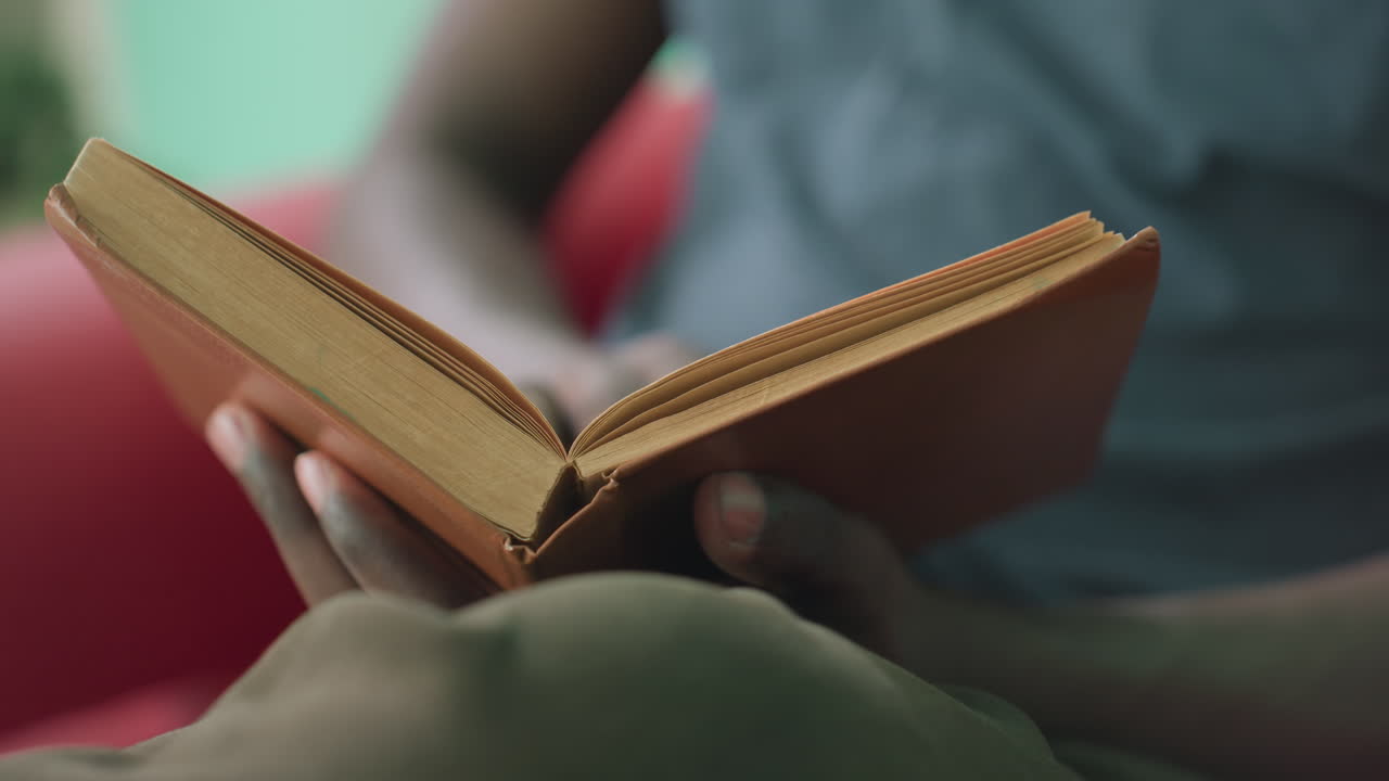 Close up of dark skin person sitting on red couch flipping page of worn brown book with careful hand while reading intently in quiet indoor setting with teal wall and warm lighting soft glow
