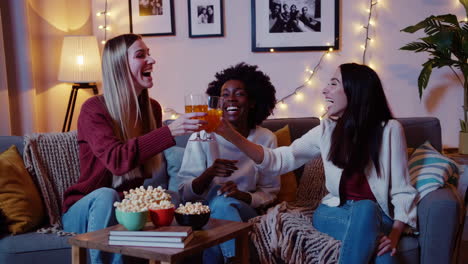 three women laughing and cheering with drinks and popcorn on a couch