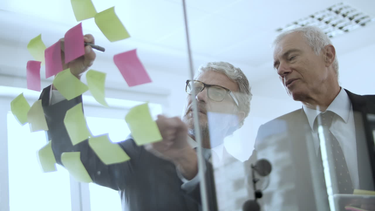 Two focused grey haired executives writing on notes on glass wall