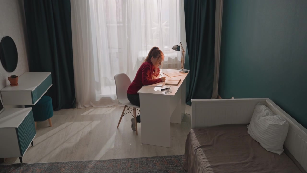 Young woman in red top is seated at white desk writing. Room features teal walls dark green curtains partially drawn. Bed with gray bedding sits nearby and various study materials